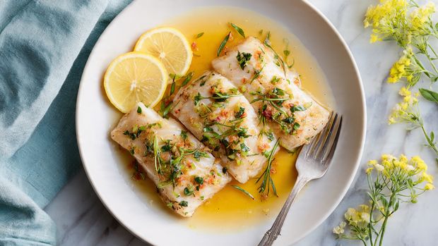 A white baking tray shows a dish with several pieces of white cooked fish placed on top of a mix of bright green asparagus pieces and vibrant red cherry tomatoes. Scattered purple olives and thin slices of cooked onion add darker color spots all around. Each fish piece is seasoned with black pepper and small green parsley leaves on top. There is a metal spatula with a wooden handle resting partly under one fish piece near the top center. A slice of lemon is visible in the top left corner outside the tray on a white marbled surface with a striped cloth nearby. The whole scene is bright and fresh-looking photo taken with an iphone --ar 4:5 --v 7