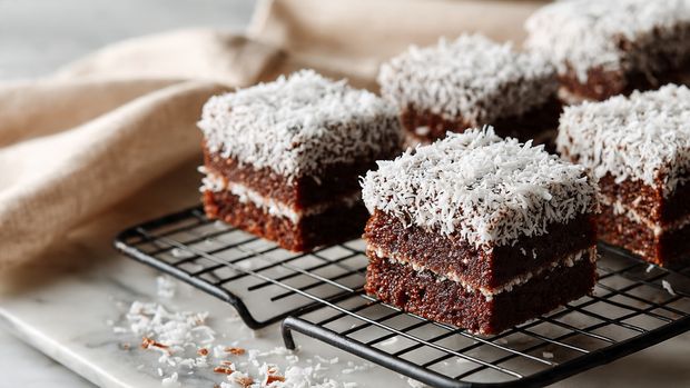 The image shows several small square chocolate cakes covered fully with shredded white coconut, giving a rough textured look. Each piece has three visible layers: a dark brown chocolate layer on the outside, a softer middle chocolate layer, and a dense cake base, all coated evenly with white coconut flakes. The squares sit on a black cooling rack over a tray with some coconut flakes scattered underneath. In the background, there is a soft cream-colored cloth, and the whole scene rests on a white marbled surface. Photo taken with an iphone --ar 4:5 --v 7