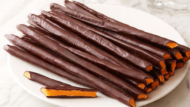 Long, thin chocolate sticks are arranged in a fan shape on a white plate with a white marbled surface underneath. Each stick is covered in smooth, dark brown chocolate on the outside and filled with a bright orange, soft center that has a slightly shiny and dense texture. Some sticks are whole, while others are broken in half to show the contrasting layers of chocolate and orange filling inside. The overall look is neat, with the dark chocolate layer wrapping tightly around the vibrant orange center, creating a strong color contrast. Photo taken with an iphone --ar 4:5 --v 7