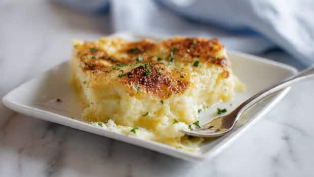 A white square plate holds a creamy yellow dish with a slightly grainy texture, topped with a golden brown crisp layer that is uneven but adds a crunchy look. Small green herb bits are sprinkled across the top, adding tiny green spots and a fresh feel. A fork rests on the right side of the plate, partly pressed into the soft dish, showing it is smooth and easy to scoop. The plate sits on a white marbled surface with a soft blue and white cloth visible in the blurred background. Photo taken with an iphone --ar 4:5 --v 7