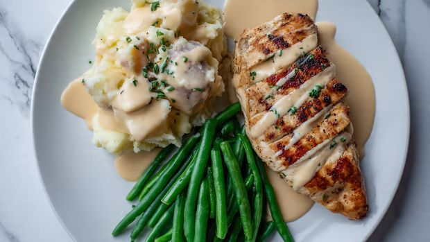 A black pan with white handles sits on a white marbled surface, holding four golden brown cooked chicken pieces. Each chicken piece is partially covered in a thick creamy light beige sauce sprinkled with small green herb bits. The chicken pieces are spread out evenly in the pan, and the sauce fills the space around them. photo taken with an iphone --ar 4:5 --v 7