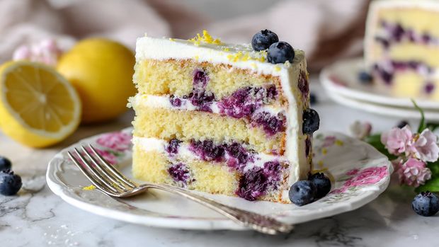 The image shows a slice of three-layer yellow cake with purple blueberries spread throughout each layer. Between each cake layer, there is a thin white cream filling. The slice sits on a white plate with a delicate pink flower pattern along the edge. A silver fork is placed next to the plate on a white marbled surface. In the background, there is a halved lemon and a white plate with more of the same cake. The overall look is bright and fresh, with soft natural lighting. photo taken with an iphone --ar 4:5 --v 7