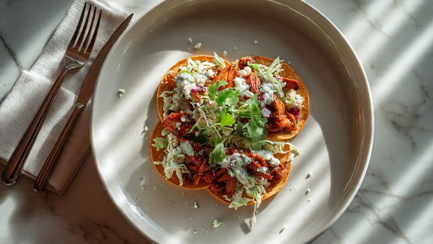 A white bowl on a white marbled surface holds a bright, fresh salad with four main layers. The bottom layer is mixed green leaves including arugula and spinach, giving a deep green color and leafy texture. On top are scattered yellow corn kernels and halved small red cherry tomatoes, adding pops of yellow and red. The next layer is golden-brown, grilled chicken pieces with herbs sprinkled on top. There are small pieces of toasted bread around the salad, some with a light spread of white dressing. White creamy dressing is drizzled over everything, with a fork and knife resting on the right edge of the bowl. A white napkin is next to the bowl with a glass of water in the corner and a bowl of white dressing with pepper specks visible on the top right edge. photo taken with an iphone --ar 4:5 --v 7