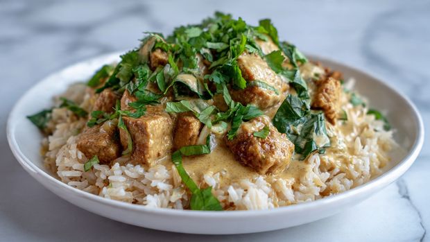 A white plate holds a dish with three main layers: the bottom layer is light brown cooked rice with soft, separated grains; on top of the rice are medium-sized browned cubes of cooked meat, showing a slightly crispy texture; the top layer has a creamy beige sauce with visible small pieces of cooked onions and green herbs, along with fresh chopped bright green leafy herbs scattered over everything. The plate sits on a white marbled surface photo taken with an iphone --ar 4:5 --v 7