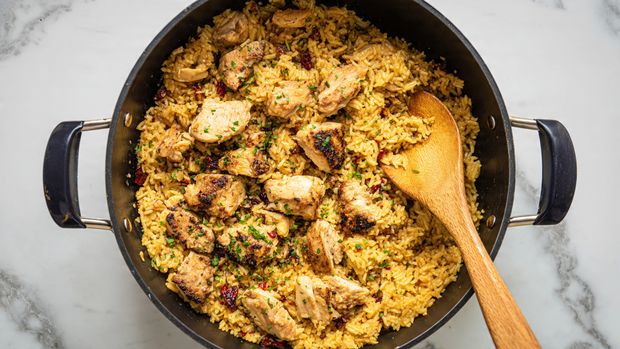 A large black pan holds a dish with two main layers: the bottom layer is cooked rice with a golden-yellow color mixed with small bits of red chili and herbs, giving a textured and moist look. On top, there are chunks of browned cooked chicken scattered evenly, each piece with a slightly charred surface showing white cooked meat inside. Small green chopped herbs are sprinkled over the chicken and rice, adding a fresh green color contrast. A wooden spoon is resting inside the pan, partially buried in the rice. The pan sits on a white marbled surface. Photo taken with an iphone --ar 4:5 --v 7