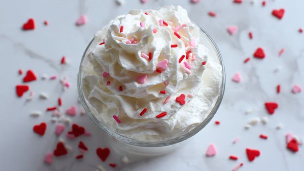 The image shows four clear measuring cups and small containers arranged on a white marbled surface. One measuring cup holds a light tan creamy liquid, and it is at the top left with a black handle. Another measuring cup with a black handle on the right contains a white liquid. Below these, there is a clear bowl filled with small, round white chips. Near the bottom center, there are five small clear packets filled with different pink and red sprinkles and candy pieces, some with white coatings. To the right of the packets, there is a small red dropper bottle with a dark liquid inside. All items are arranged neatly with a bright, clean look photo taken with an iphone --ar 4:5 --v 7