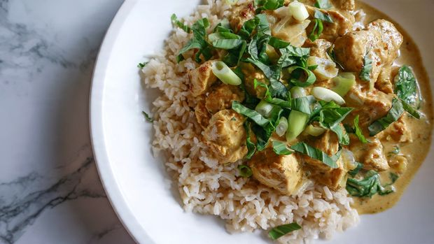 A close-up view of a skillet filled with a creamy white sauce that has a smooth texture with small bubbles on the surface. In the sauce are pieces of browned, small, irregular chunks of meat spread evenly throughout. Bright green spinach leaves are mixed in, some slightly wilted, along with finely chopped light beige onions scattered around. The skillet has a dark surface, with a white marbled background visible at the edges. photo taken with an iphone --ar 4:5 --v 7
