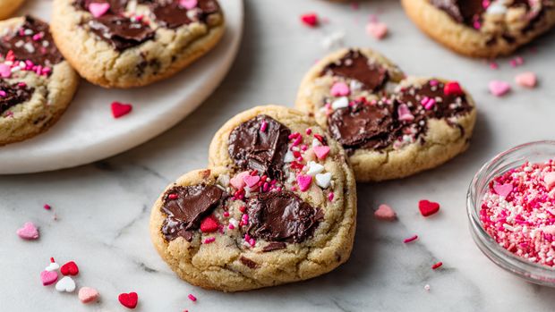 A plate filled with many heart-shaped cookies sits on white parchment. Each cookie is light brown with dark chocolate chunks embedded inside and on top. Small red, pink, and white heart-shaped sprinkles are scattered across the cookies, adding bright spots of color. The cookies have a slightly rough texture with visible sugar grains and chocolate melting in places. Around the plate are clear lids and a small transparent bowl filled with more red, pink, and white sprinkles, all placed on a white marbled surface. Photo taken with an iphone --ar 4:5 --v 7