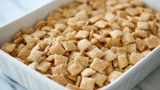 Pistachio Puppy Chow Recipe 4 The image shows a close-up of a white baking dish filled with a single layer of light golden, square-shaped cereal pieces that have a textured, woven pattern. The pieces look slightly shiny and crunchy, with some small clusters scattered throughout. The dish sits on a white marbled surface, and the focus is on the top of the cereal layer, highlighting its crisp texture. photo taken with an iphone --ar 4:5 --v 7