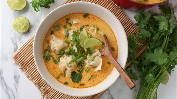 A black pot with a red outer edge filled with creamy orange soup showing hints of oil on the surface, with several light beige dumplings floating inside. Small green herbs, possibly chopped scallions, and dark slices, likely mushrooms, are scattered throughout. A utensil with a wooden handle and metal scoop is partially submerged in the soup. The pot rests on a white marbled surface with a white cloth that has black geometric patterns underneath. Photo taken with an iphone --ar 4:5 --v 7