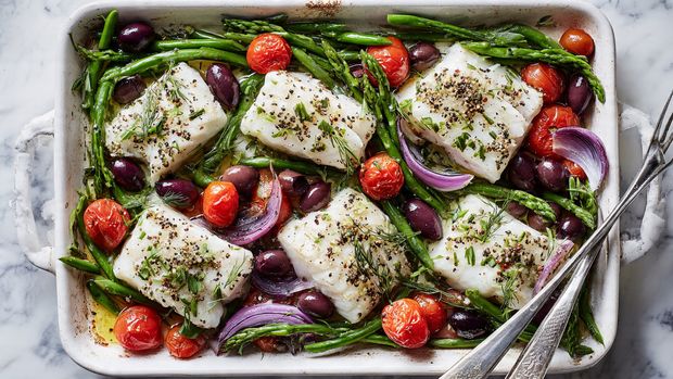 The image shows a white rectangular baking tray filled with six pieces of white fish fillets placed evenly across the tray. Around and between the fish pieces are bright green asparagus cut into small sections, whole red cherry tomatoes, thin slices of purple onion, and dark purple olives. The fish fillets are sprinkled with black pepper and drizzled with a little olive oil. The tray is on a white marbled surface, and a pair of silver tongs rests on the bottom right corner of the tray. photo taken with an iphone --ar 4:5 --v 7